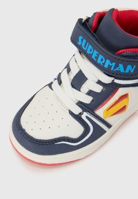 Baskets montantes en bleu marine et blanc, affichant le logo "SUPERMAN", des accents rouges et des lacets blancs, inclinées pour mettre en avant les détails de conception.