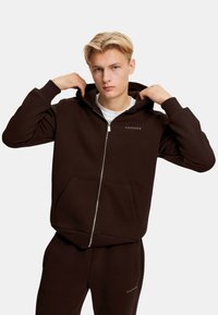 Mørkebrun zip-up hettegenser laget av mykt stoff, med en front lommesekk, ribbede mansjetter og hette. Liten logo på brystet.