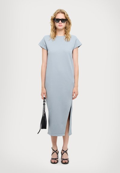 Vestido midi azul claro de tela suave con mangas cortas, abertura lateral y sandalias planas. Los accesorios incluyen un pequeño bolso negro.