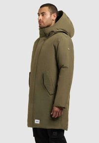 Olive drzewny parka z dużym kapturem, bocznymi kieszeniami, gładką fakturą i białą metką materiałową z przodu na dole.