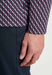 Marineblauw shirt met een geometrisch rood en wit patroon. Close-up van een hand die op donker marineblauwe broek rust, waarbij de textuur en pasvorm van de stof worden getoond.