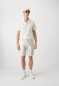 Hvit kortermet skjorte med et strukturert mønster og brystlomme, kombinert med lyse shorts og hvite joggesko. Tilbehør som solbriller og caps.