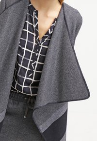 Grijze cardigan met een open voorkant, voorzien van een gebreide textuur. Daaronder een marineblauwe overhemd met knopen en een wit ruitpatroon.