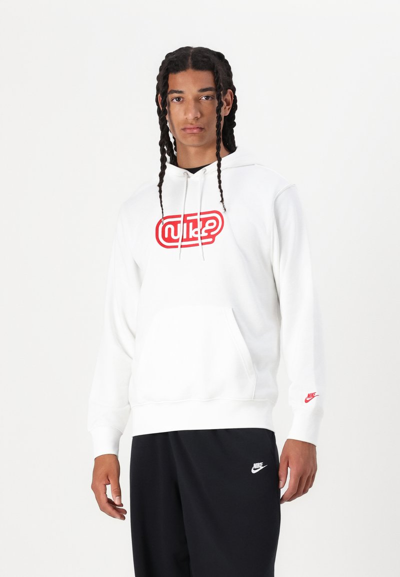 Felpa bianca in cotone con logo rosso "Nike", tasca a marsupio frontale, cappuccio con coulisse e polsini a costine. Abbinata a pantaloni scuri.