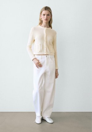 Femme debout, portant un cardigan en maille crème à boutons, un pantalon blanc à jambes larges, des chaussures blanches et des bagues dorées à une main.