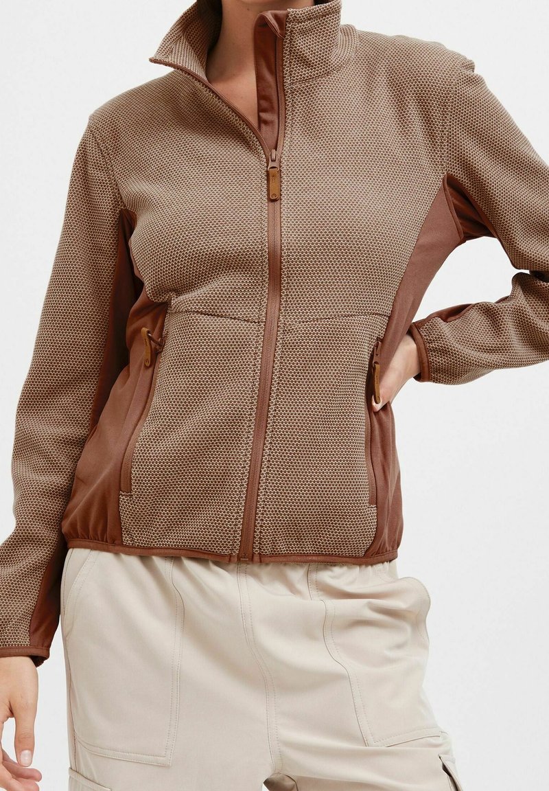 Femme portant une veste marron texturée à fermeture éclair avec poches latérales et un pantalon crème, debout, une main sur la hanche et l'autre relâchée.