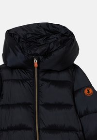 Zwarte gewatteerde pufferjas met grote capuchon, voorrits met bruine trekker, en ronde bruine logo patch op de mouw.