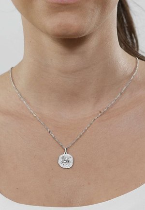 Silberne Halskette mit quadratischem Anhänger, der ein geprägtes Widder-Motiv zeigt, getragen am Hals einer Frau über einem weißen Oberteil.