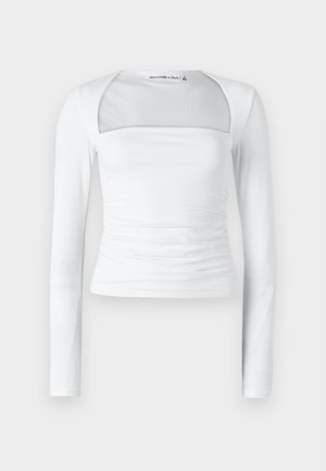 Valge pikkade varrukatega topp ruudukujulise kaelusega, millel on läbipaistev paneel, volditud külgelemendid ja sile veniv kangas.