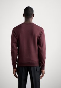 Uomo in piedi con le spalle alla macchina fotografica, indossando una felpa a maniche lunghe bordeaux e pantaloni neri, con uno sfondo chiaro e neutro.