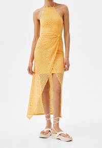 Gul virkade klänning med halterneck, slits till mitten av låret och omslagdesign; bärs över en åtsittande gul underklänning. Ett par kraftiga vita sandaler.