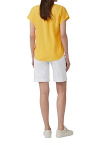 T-shirt en lin jaune à manches courtes, ourlet arrondi, associé à un short en denim blanc avec des poignets roulés et des baskets de couleur claire.