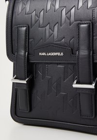 Borsa in pelle nera con motivo geometrico in rilievo, hardware argentato e spallacci regolabili. Marchio visibile in modo prominente sul davanti.