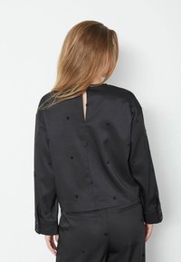 Blusa nera, textured con apertura a chiave sul retro, maniche lunghe e punti decorativi su tutta la superficie. Vestibilità sartoriale con una finitura liscia.