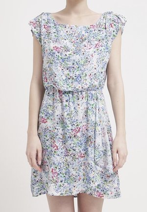 Vrouw draagt een mouwloze, knie-lange jurk met een blauw, roze en groen bloemenpatroon en een gestrikte riem aan de rechterkant.