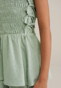 Šviesiai žalia suknelė su susmulkintu korpusu, banguotomis juostelėmis ir tekstūruotu audiniu, pasižyminčiu mažomis, iškilusiomis taškelių. Platėjanti sijonas tęsiasi žemiau juosmens.