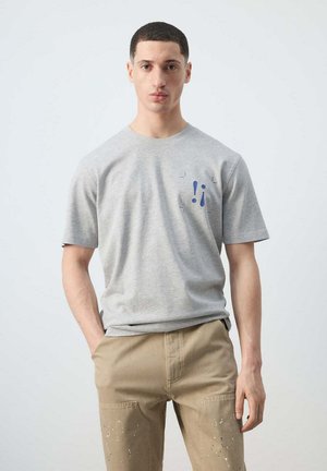 Joven con camiseta gris con estampado azul de signos de puntuación y pantalones beige con salpicaduras de pintura, de pie con una mano en el bolsillo.