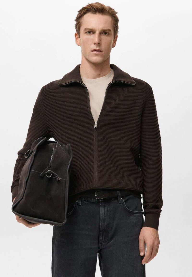 Brązowy sweter na zamek z dzianiny o fakturowanym wykończeniu, noszony na beżowej koszuli. W lewej ręce trzymane są czarne dżinsy oraz ciemna zamszowa torba.