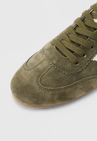 Κοντινό πλάνο του μπροστινού μέρους αθλητικού παπουτσιού από σουέτ σε πράσινο ελιάς με κορδόνια στο ίδιο χρώμα και λεπτομερή ραφή πάνω σε ανοιχτό φόντο.