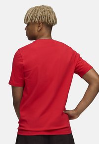 Rød bomuld T-shirt med rund halsudskæring og korte ærmer; model set fra bagenden, iført sorte bukser og en sølvkæde halskæde.