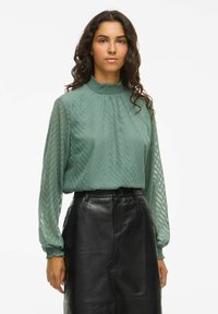 Blusa verde con texture e motivo a zigzag, colletto alto e maniche lunghe, abbinata a una gonna di pelle nera. Finitura liscia e vestibilità rilassata.