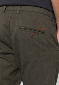 Donkergroene broek met een gestructureerd patroon, voorzien van een knoopsluiting op de achterzak en een klein leren accentlabel.