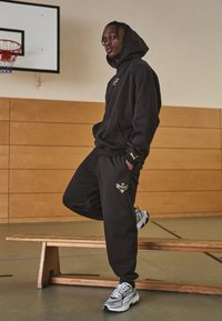 Black hoodie with gold logo, matching sweatpants featuring gold emblem. Light gray athletic shoes with dark accents. Wooden bench in background.