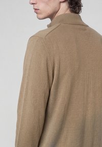 Jeune homme portant un pull en maille marron clair avec un col montant côtelé, vu de dos et de profil sur fond uni.