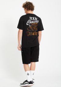 T-shirt en coton noir avec un texte graphique blanc et orange au dos, associé à un short noir. Le modèle porte des chaussettes blanches et des baskets noires.