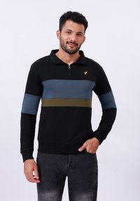 Černá mikina se zipem, s modrými a olivově zelenými vodorovnými pruhy. Má stojáček a malé zlaté logo na hrudi.