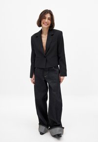 Moteris šypsosi, vilkinti juodą trumpą švarką su viena saga ir dideles, plačias juodas džinsus su sukniuotais kraštais.