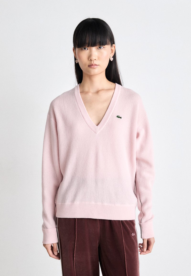 Pull V-neck rose clair en matière douce avec des manches longues et des poignets côtelés. Logo Lacoste sur la poitrine gauche. Accompagné d'un pantalon bordeaux en velours.