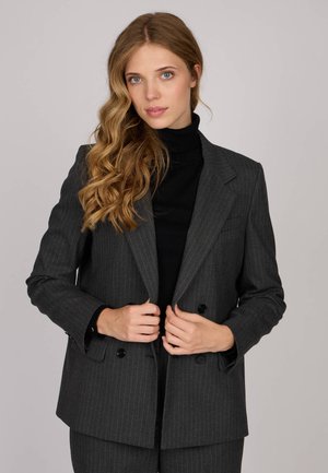 Blazer gris à rayures avec un design à double boutonnage, revers crantés et boutons noirs. Porté par-dessus un pull noir à col roulé.