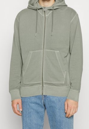 Mężczyzna noszący jasnooliwkową bluzę z kapturem zapinaną na zamek, z przednimi kieszeniami i sznurkami, w połączeniu z niebieskimi dżinsami.