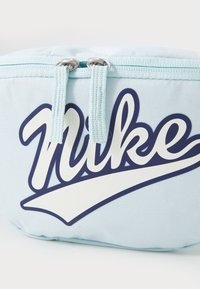 Šviesiai mėlynas audinio maišelis su išraiškingu tamsiai mėlynu Nike logotipu, dviem užtrauktuko trauklėmis ir lygiu paviršiumi su subtiliu raštu.