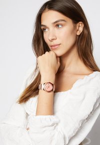 Montre en or rose avec un cadran rond et un fond rose, arborant des index des heures argentés et un bracelet en silicone rayé. Portée au poignet sur un tissu blanc.
