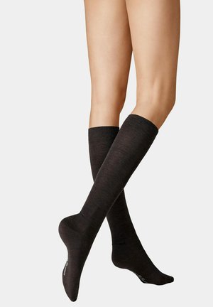 Crossed legs wearing black knee-high socks with visible brand text on the sole against a plain light background.