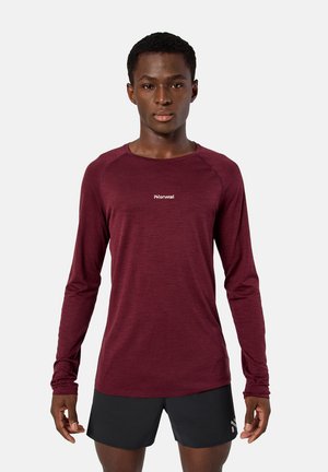 Joven vestido con una camiseta deportiva de manga larga color burdeos con el logo "Norwal" y pantalones cortos negros, posando contra un fondo claro y sencillo.