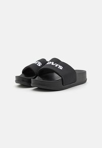 Sandales noires à semelle plate avec de larges sangles en tissu présentant un texte logo blanc, une semelle en caoutchouc texturé et un design ouvert au niveau des orteils.