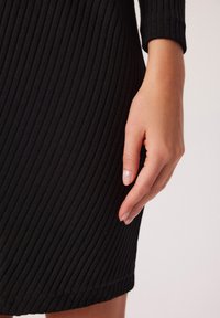 Czarna, żebrowana sukienka o dopasowanym kroju. Zbliżenie na dłoń opartą na dolnej krawędzi, ukazujące teksturę i elastyczność materiału.