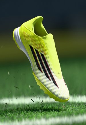 Helle gelbe Fußballschuhe mit einer strukturierten Oberseite, die schwarze und rote diagonale Streifen aufweist, und einer weißen Sohle. Hintergrund aus grünem Gras.