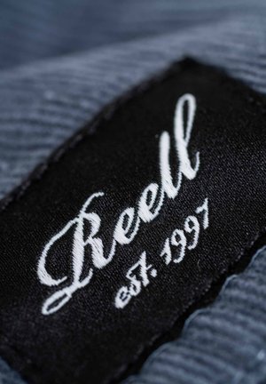 Black fabric tag with white stitched text "Reell" and "est. 1997," set against a textured blue background.