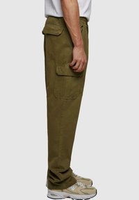 Olivenfarvede cargo bukser med sidelommer, elastisk talje, tapered ben og en glat tekstur, parret med beige sportssko.