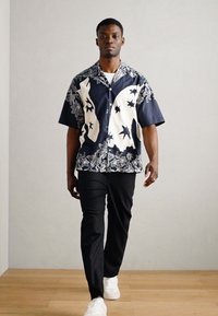 Κοντομάνικο πουκάμισο ναυτικού χρώματος με λευκά λουλούδια και αστέρια, συνδυασμένο με μαύρο παντελόνι και λευκά αθλητικά παπούτσια. Απαλή, ελαφριά ύφανση.