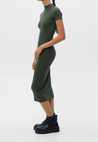 Enganliegendes, kurzärmeliges, dunkelgrünes Kleid mit hohem Kragen, knielangem Saum, weichem Stoff und seitlichen Taschen; kombiniert mit schwarzen Schnürboots.