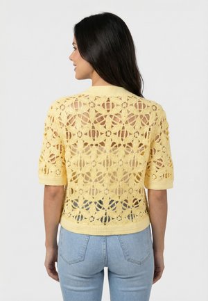 Femme de dos portant un haut jaune en crochet à manches courtes avec des motifs floraux sur un jean bleu clair.