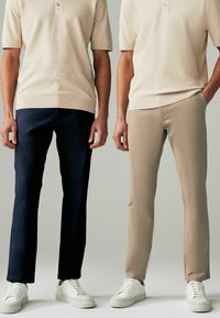 Ein Paar Herren-Hosen: eine dunkelblau und eine beige, beide mit glatter Textur, geradem Schnitt und getragen mit weißen Sneakers.