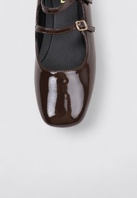 Zapato Mary Jane de charol marrón con dos correas ajustables, punta cuadrada y textura suave, situado sobre un fondo gris claro.