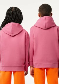 Due persone che indossano felpe rosa abbinate con una finitura texturizzata. Design con cappuccio, cuciture visibili, abbinate a pantaloni sportivi arancioni.
