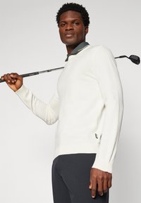 Maglione a maglia grigio chiaro con scollo rotondo, maniche lunghe e dettagli a costine. Una mazza da golf poggia sulla spalla, completando il look informale.